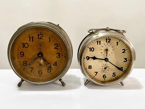 Two vintage silver-colored alarm clocks with worn faces displayed on a white surface against a plain background