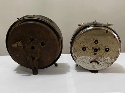 Back view of two vintage silver-colored old-fashioned alarm clocks on a white surface with worn details